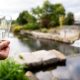 Hand hält eine Filament-Glühbirne vor einem Wehr an einem Fluss; Wasserlauf, Ufer und Bäume im Hintergrund unscharf.