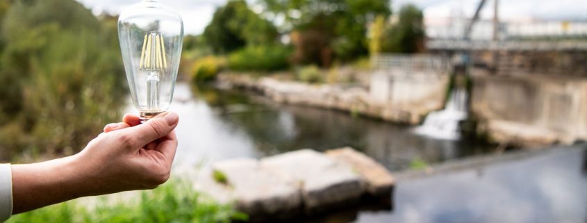 Hand hält eine Filament-Glühbirne vor einem Wehr an einem Fluss; Wasserlauf, Ufer und Bäume im Hintergrund unscharf.